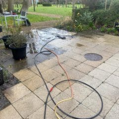 A tiled patio with garden hose pipes lying across wet and dry tiles hints at a recent driveway jet wash. Potted plants line the edge, while garden chairs and a table sit in the background near a grassy lawn with trees and shrubs.