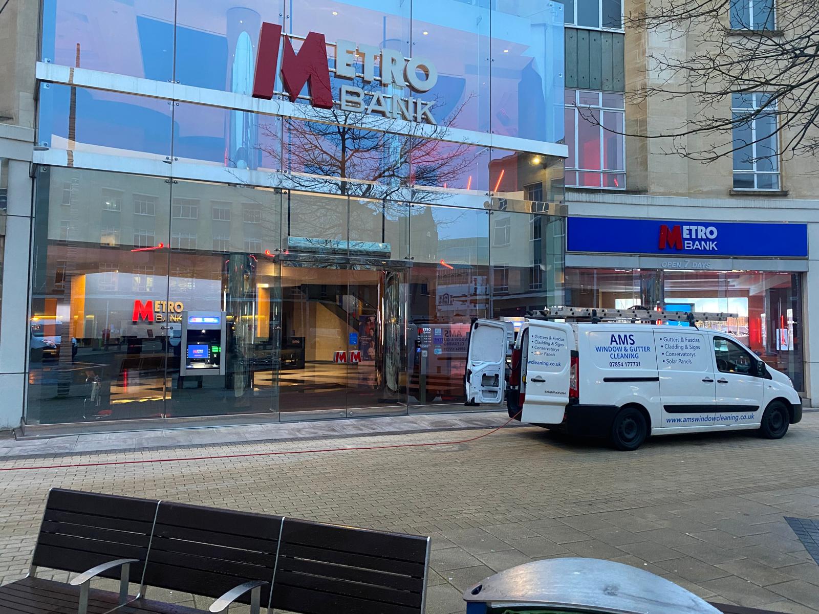 A white AMS van is parked outside a Metro Bank branch with its back door open and cleaning equipment inside. The bank’s glass facade and ATMs are visible, with reflections of nearby buildings on the glass.