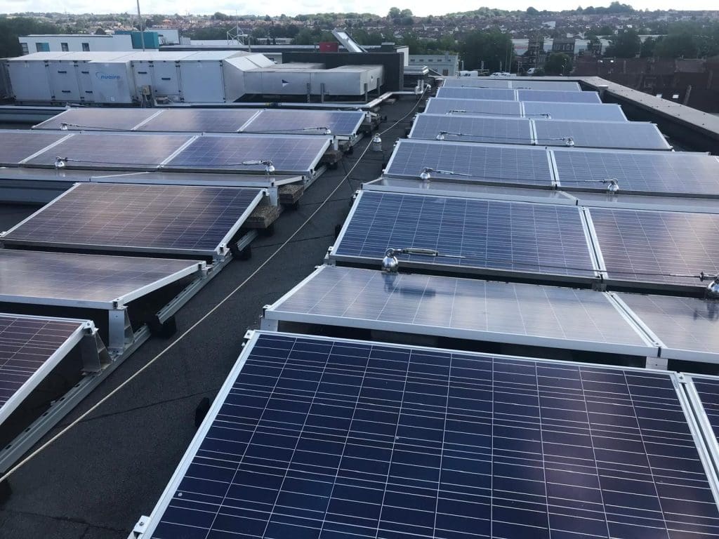 Rows of solar panels are installed on a flat rooftop, with buildings and a residential area visible in the background under a cloudy sky.